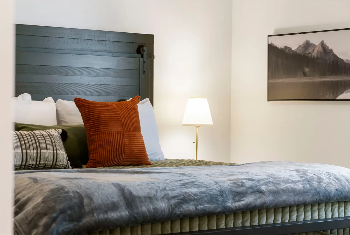 An inviting bedroom with a king-size bed dressed in textured linens and accented by a bright orange pillow. A bedside lamp emits a warm glow, and a framed mountain landscape adds a touch of sophistication and peace to the space.