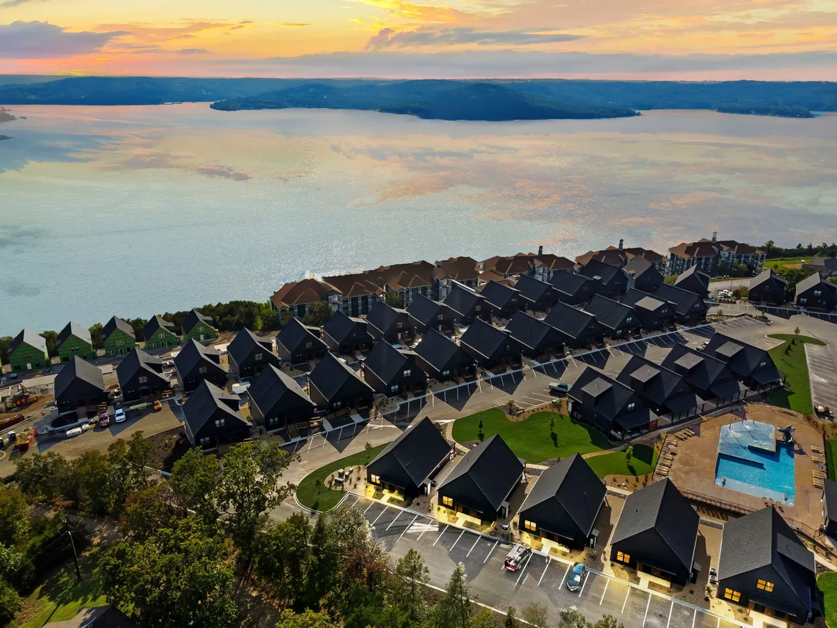A wide exterior view of the lake, rooftops of houses, a swimming pool, and landscaped areas. The scene is serene, featuring sunset views, spaces for relaxing by the lake, and opportunities for family gatherings and shoreline strolls.