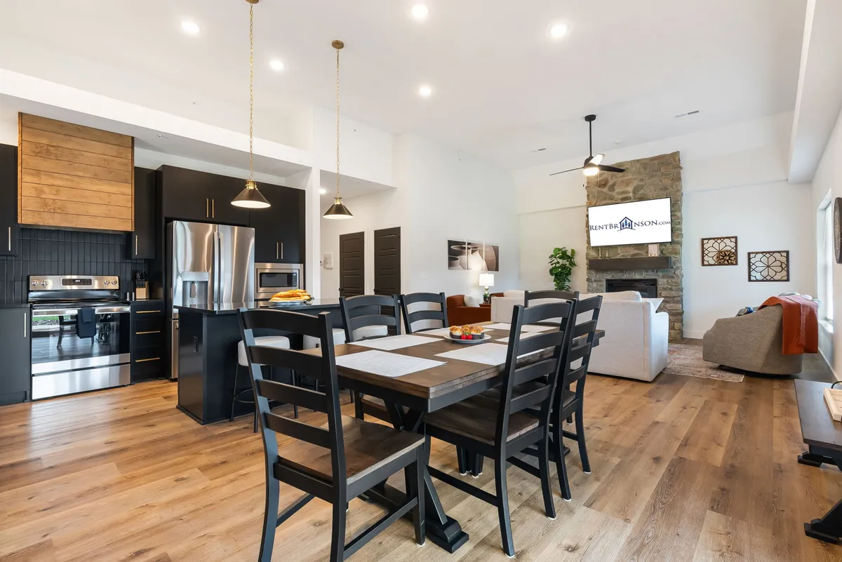 This wide-angle shot captures the open-concept layout of the kitchen, dining area, and living room. The spacious kitchen island, large dining table, and comfortable living area with a prominent stone fireplace are all visible. Large windows provide ample natural light, and the setup encourages communal gathering and easy movement throughout the shared space.