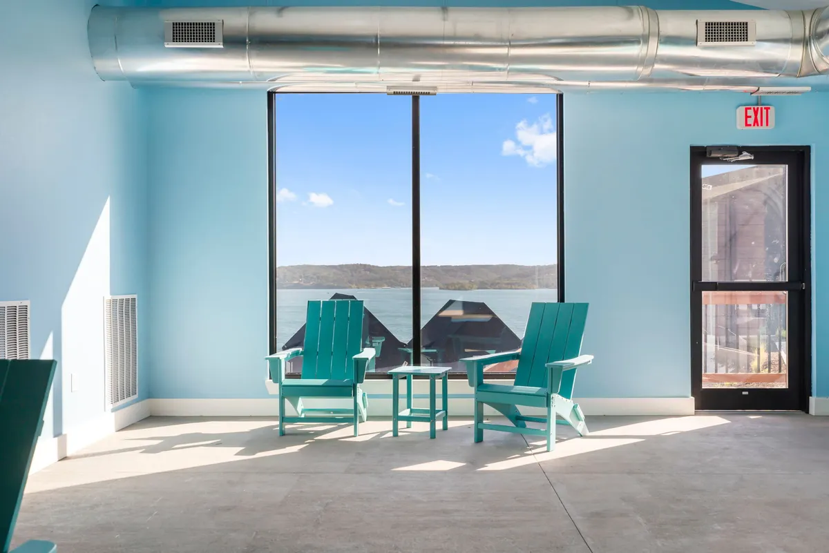 A bright, tranquil room featuring large windows with a view of the lake, two teal Adirondack chairs, and a small table—perfect for enjoying coffee while watching the water. The scene evokes peace and inspiration, ideal for starting the day or taking a restorative break.