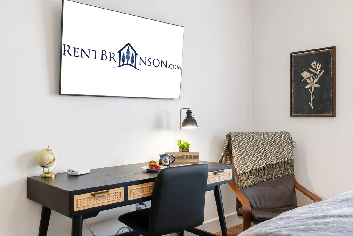 A compact but inviting workspace setup, featuring a desk decorated with stylish elements, a comfortable chair, a TV with branding, and framed wall art. The area feels cozy and conducive to productivity.