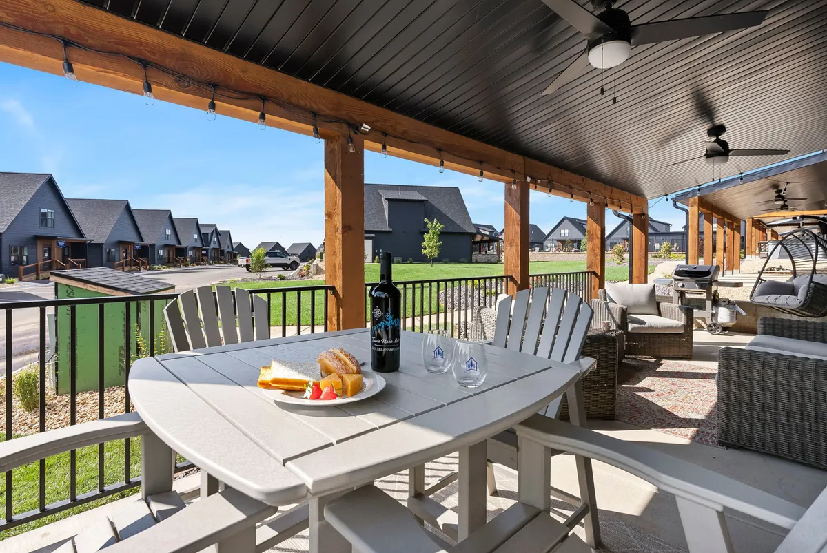The image features an outdoor dining table set with food and drinks, surrounded by comfortable seating and beautiful landscaping. The view includes neighboring houses, reinforcing a sense of community. The mood is relaxed and social, ideal for gatherings or quiet moments alike.
