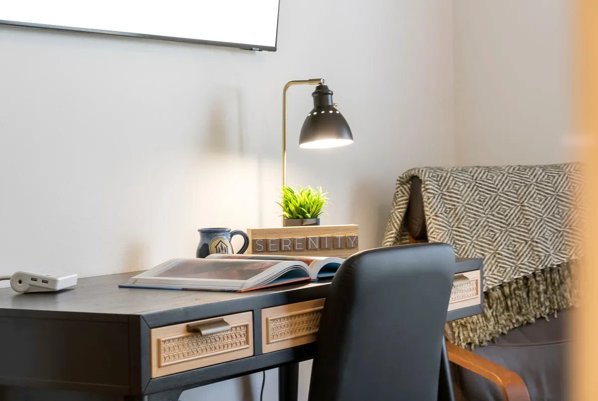A stylish desk setup with decorative elements, a cozy lamp, a green plant, an open book, and a mug. The serene workspace radiates calm and productivity, making it easy to imagine settling in for a focused remote work session.