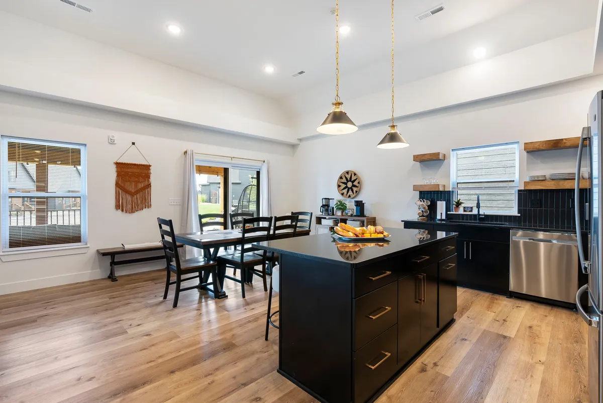 A bright kitchen view with a black island, adjacent dining table, abundant natural light streaming through large windows, and tasteful decorative touches. The kitchen appliances and layout are practical and welcoming, ideal for both cooking and gathering.