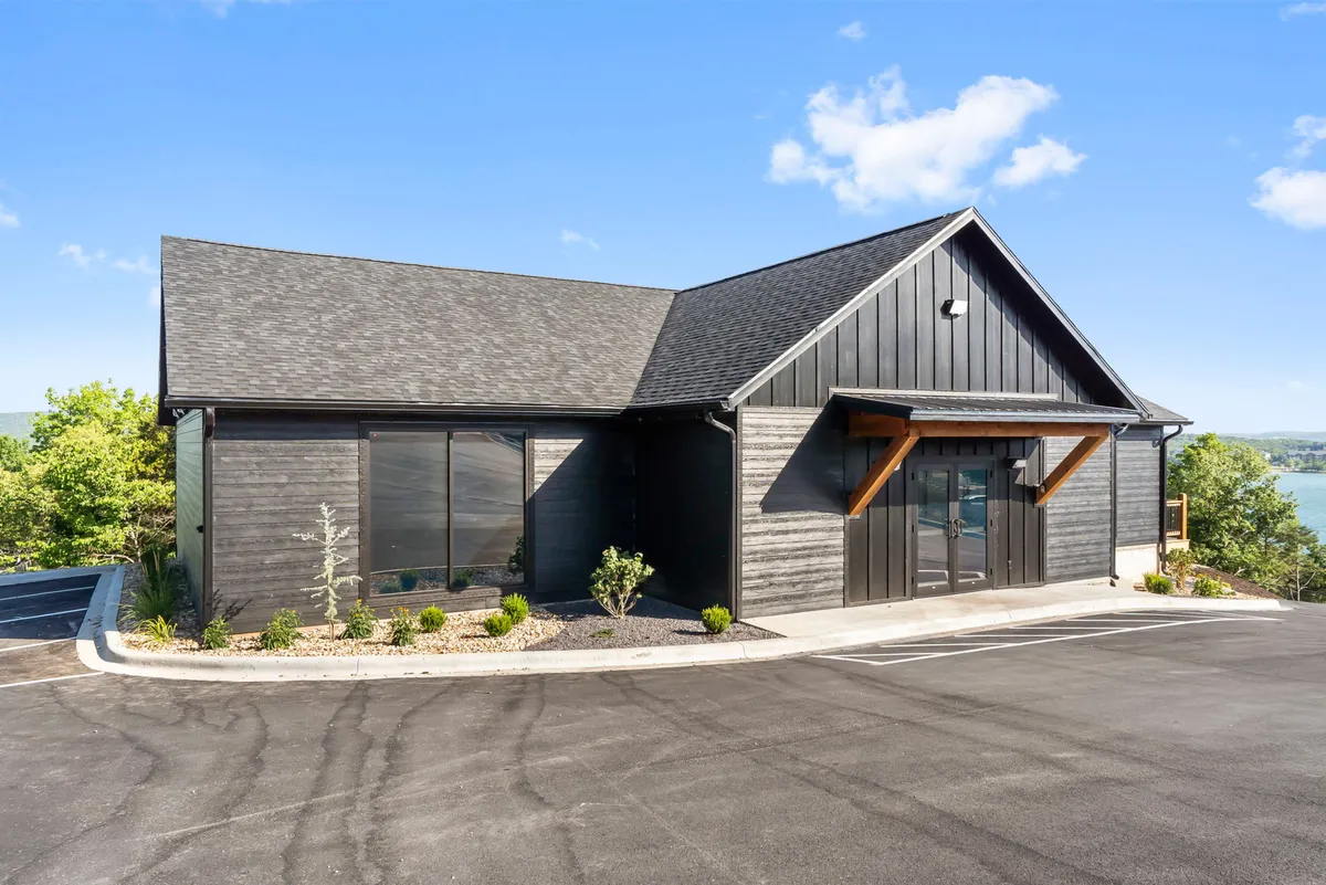 An exterior shot of a modern building with large windows and wooden accents, surrounded by a landscaped garden. The scene implies a welcoming environment for socializing, with outdoor seating areas and attractive, natural surroundings.