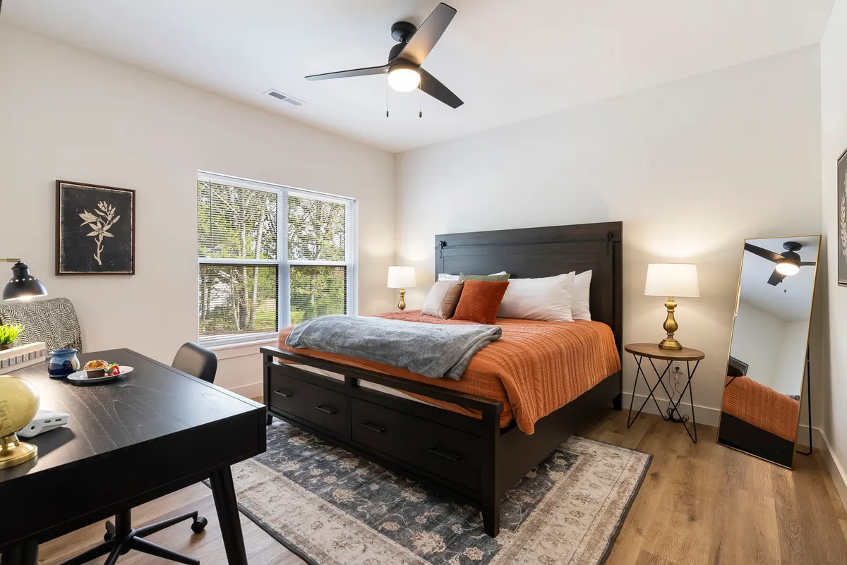 A stylish bedroom features a cozy bed with an orange throw blanket, a desk and chair set up next to a large window that bathes the room in natural light. A decorative mirror and wall art add to the inviting vibe, making the workspace feel both productive and homey.