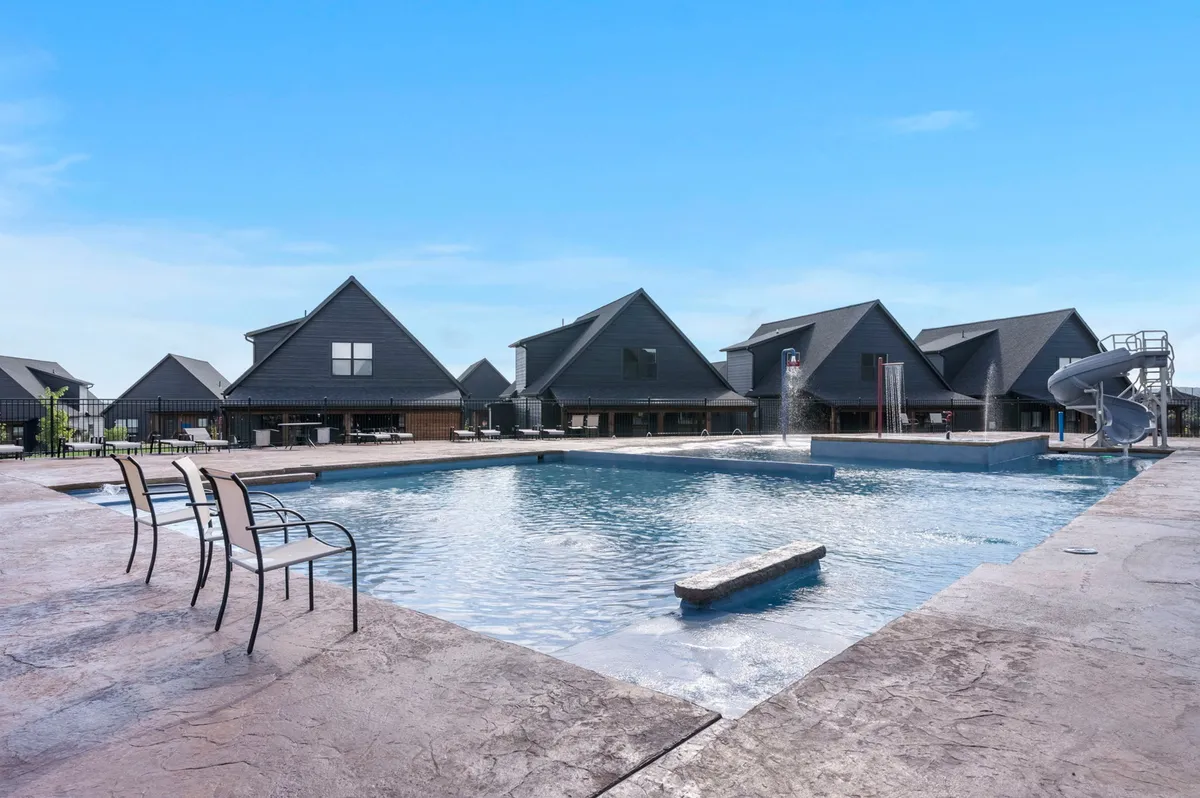 A vibrant outdoor pool scene with a water slide, pool deck, and surrounding community buildings. The mood is cheerful, with open space for sunbathing, swimming, and family fun, capturing the essence of resort-style community living.